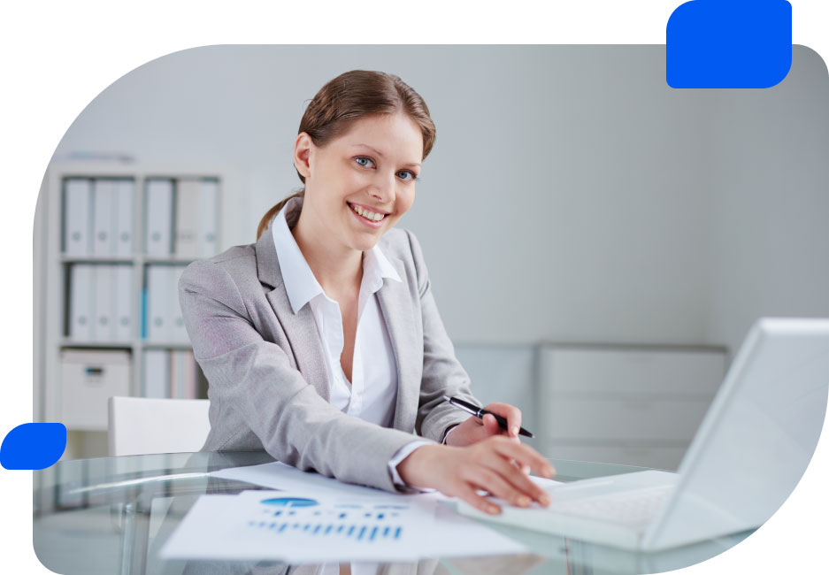 Smiling businesswoman sitting at a desk with a laptop and charts, looking engaged and professional in an office setting.